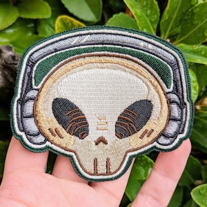 May include: An embroidered patch featuring an alien skull wearing headphones. The skull is green, grey and brown with large black eyes and a detailed design.