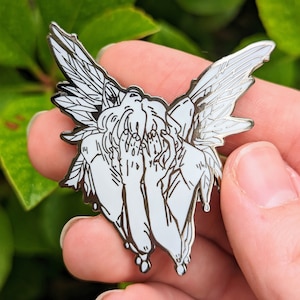 May include: A silver-toned enamel pin depicting a weeping angel with wings outstretched. The angel is depicted with its face hidden in its hands, and tears are dripping from its eyes.