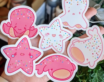May include: Six colorful stickers featuring various sea creatures with a sprinkle pattern. The stickers include a pink octopus, a white starfish, a pink crab, a white dolphin, a white jellyfish, and a pink starfish.
