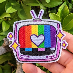 May include: A rainbow-colored television screen with a white heart in the center. The television is surrounded by a purple frame with yellow stars on the sides. The television is a patch.