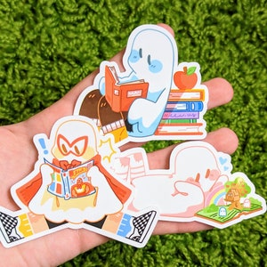Puede incluir: Tres pegatinas de fantasmas coloridos con diferentes expresiones. Un fantasma está leyendo un libro, otro está leyendo un cómic y el tercero está acostado en un libro con un arcoíris y una casa en el árbol.