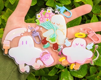 May include: Four colorful ghost-shaped keychains with different designs. One ghost holds a bouquet of pink flowers, another carries a paintbrush and a bucket of hearts, a third has a halo and wings, and the last one is holding an arrow.
