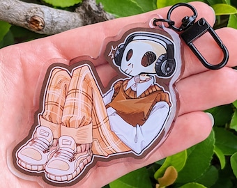May include: A clear acrylic keychain featuring a cartoon skeleton wearing a brown sweater vest, plaid pants, and white sneakers. The skeleton is wearing headphones and has a surprised expression.