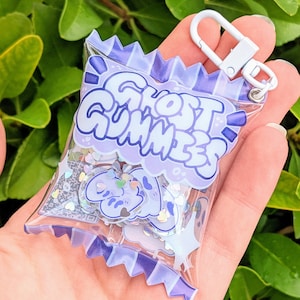 Peut inclure: Un porte-clés en forme de sac en plastique transparent avec un fermoir blanc, rempli de confettis et de breloques en forme de fantômes. Le sac porte l'inscription "Ghost Gummies" dans un motif de nuage violet. Le porte-clés est tenu dans une main.
