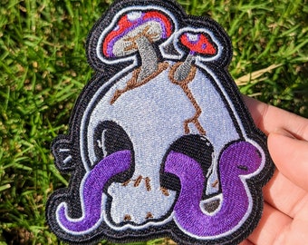 May include: A black and white embroidered patch of a skull with two red and white mushrooms growing out of the top. Two purple snakes are wrapped around the skull.