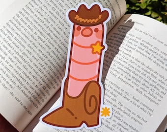 Puede incluir: Un personaje de dibujos animados rosa y marrón que lleva un sombrero de vaquero y botas. El personaje es un gusano antropomórfico largo, delgado, con una estrella en el pecho. El personaje está de pie sobre un libro con el texto "All About Pies" visible.