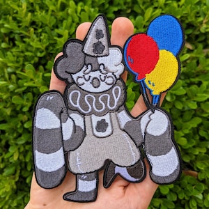 May include: An embroidered patch depicting a clown holding three balloons. The balloons are red, blue and yellow. The clown is wearing a hat and has a sad expression.