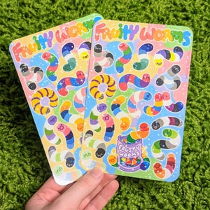May include: Two sheets of colorful stickers featuring gummy worms with rainbow colors and glitter. The stickers say "Fruity Worms" and feature a cartoon gummy worm bucket.
