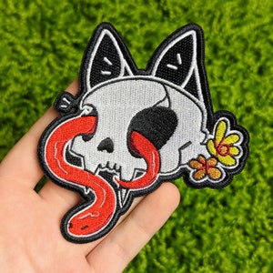 Könnte beinhalten: Gestickter Bügel-Patch mit einem Cartoon-Schädel mit herausgestreckter roter Zunge, der eine schwarze Katzenmaske trägt und eine gelbe Blume dahinter hat.