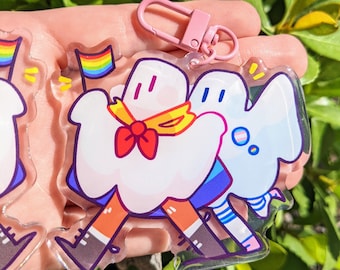 May include: A clear acrylic keychain featuring two cartoon ghosts holding a rainbow flag. The ghosts are wearing colorful clothing and have happy expressions.