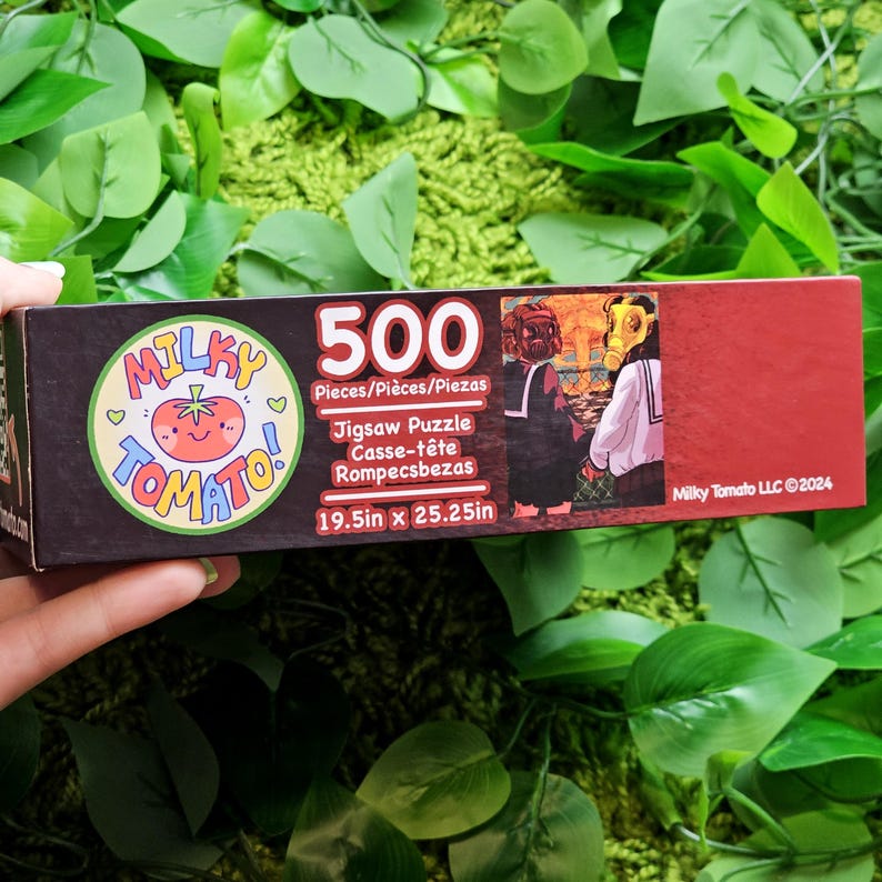 May include: A red box with a 500 piece jigsaw puzzle of two people in school uniforms standing in front of a mushroom cloud. The box has a logo for Milky Tomato LLC and the text "500 Pieces/Pi&egrave;ces/Piezas Jigsaw Puzzle Casse-t&ecirc;te Rompecsbezas 19.5in x 25.25in Milky Tomato LLC &copy;2024".