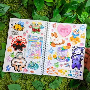 May include: An open notebook with colorful stickers featuring cartoon characters, food, and text. The stickers include a bunny, a worm, a frog, a mushroom, and a creature from the black lagoon. The notebook has a spiral binding and is open to a page with the text "Kinda Fruity" and "Fruity Worms".