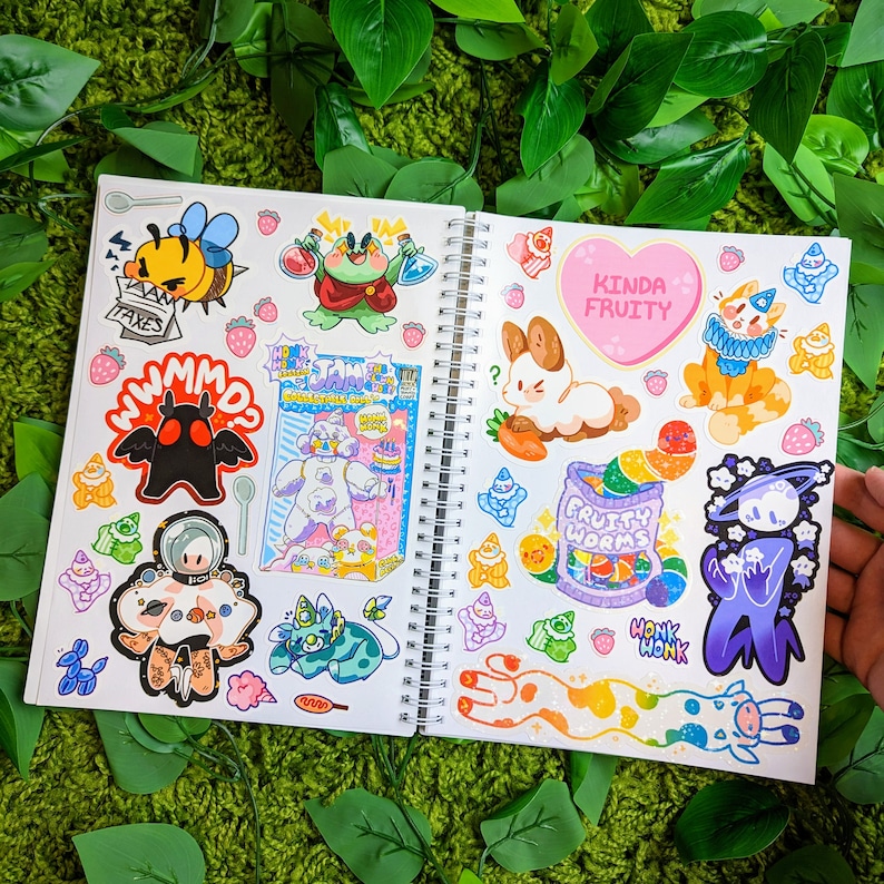 May include: An open notebook with colorful stickers featuring cartoon characters, food, and text. The stickers include a bunny, a worm, a frog, a mushroom, and a creature from the black lagoon. The notebook has a spiral binding and is open to a page with the text "Kinda Fruity" and "Fruity Worms".