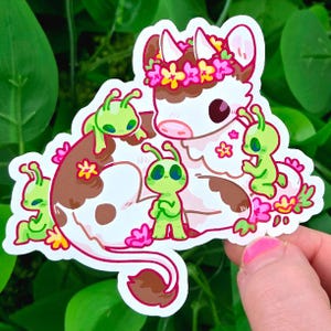 May include: A whimsical sticker featuring a cartoon cow with brown and white spots, adorned with a floral crown. The cow is surrounded by green alien figures and pink flowers, creating a playful and imaginative design.