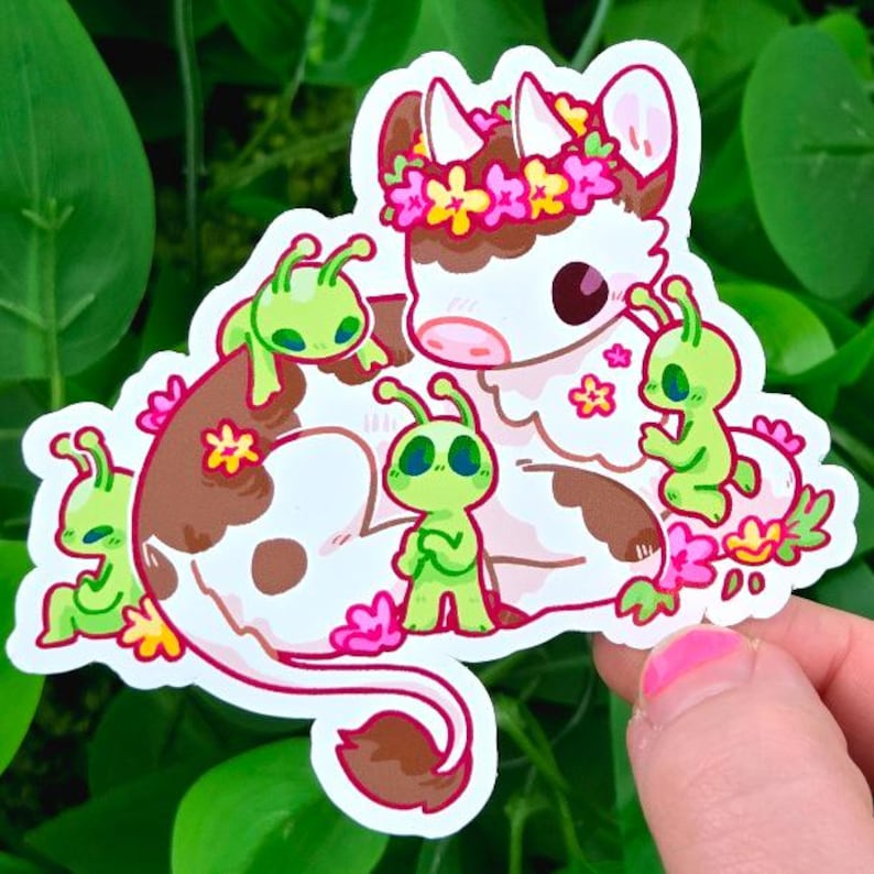 May include: A whimsical sticker featuring a cartoon cow with brown and white spots, adorned with a floral crown. The cow is surrounded by green alien figures and pink flowers, creating a playful and imaginative design.
