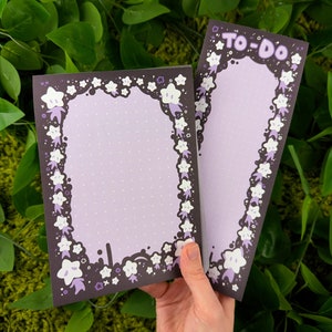 May include: Two purple and white patterned to-do list notepads with a black background and a white star design. The notepad on the left has a dotted background and the notepad on the right has a solid background. The notepad on the right has the text "TO-DO" in white letters.