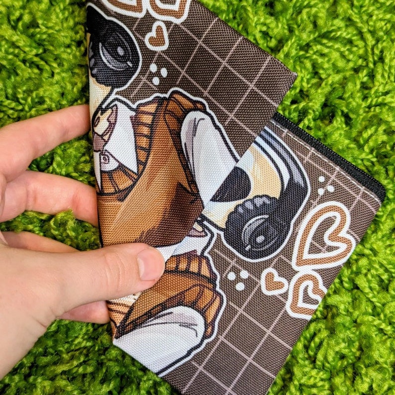 May include: A brown zippered pouch with a white and brown cartoon character design. The character is wearing a brown sweater vest and headphones. The pouch has a grid pattern and hearts.