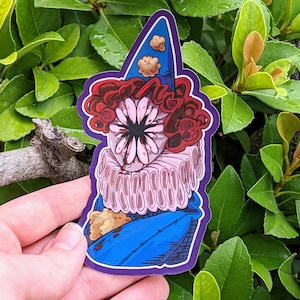 May include: A sticker of a clown with a large, pink flower for a face. The clown is wearing a blue and white hat with a red and white ruffle collar. The clown is holding a small, brown teddy bear.