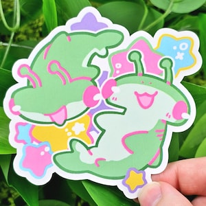 May include: A sticker featuring two cartoon alien characters in shades of green, pink, and purple. The aliens have large eyes and antennae, with a smiling expression. Surrounding the aliens are colorful stars and shapes.