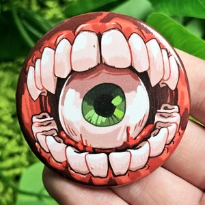 Puede incluir: Una boca roja carnosa con dientes afilados, sosteniendo un gran globo ocular verde. El globo ocular está rodeado de sangre y la boca está abierta de par en par.