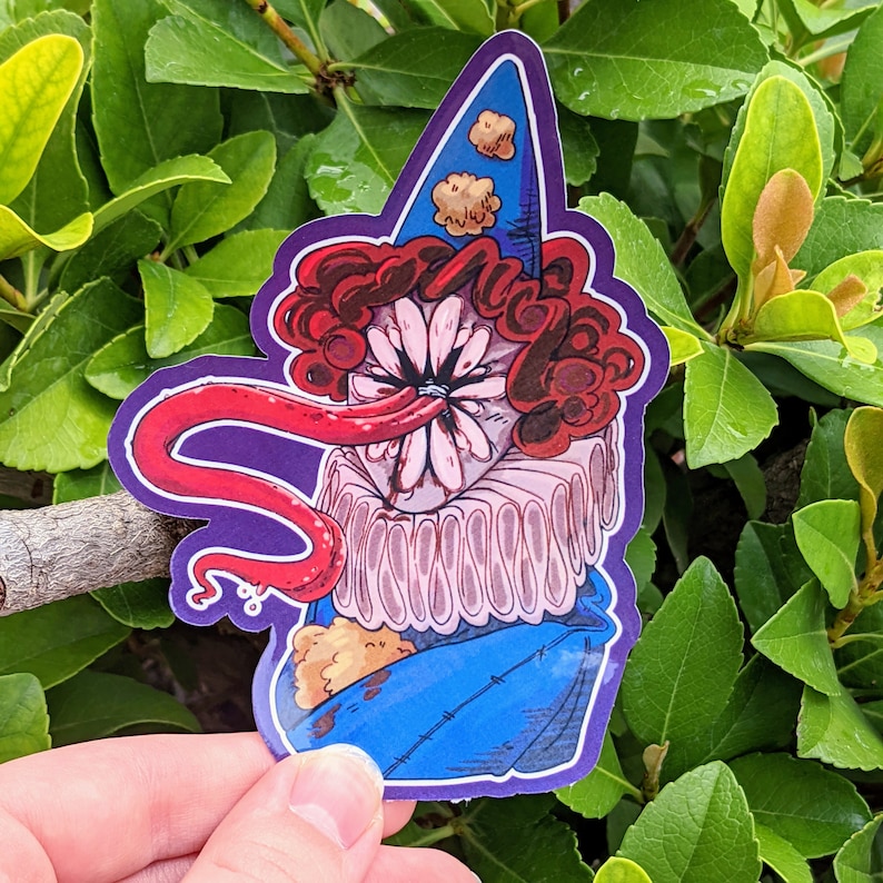May include: A sticker of a cartoon character with a red tongue, red hair, and a white flower-like face. The character is wearing a blue and white jester hat and a pink ruffled collar. The sticker is on a green leafy background.