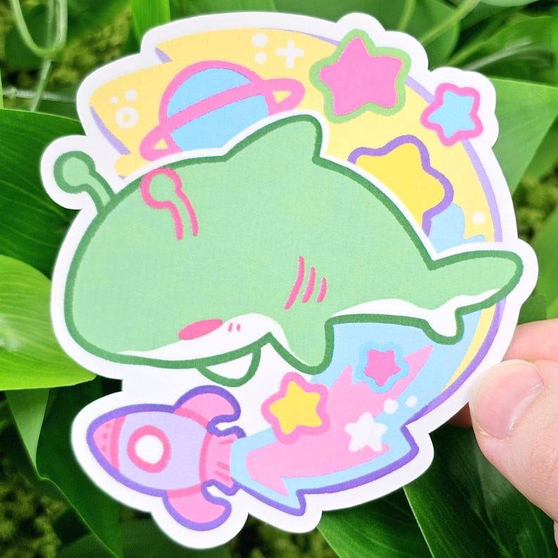 May include: A die-cut sticker featuring a green shark with pink accents, surrounded by a space-themed design. The design includes a rocket, planets, stars, and a yellow and blue background. The sticker is held against a background of green leaves.