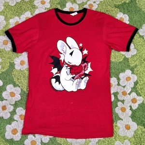 May include: A red ringer t-shirt with black trim on the collar and sleeves. The center graphic features a white creature with black bat wings holding a red heart, surrounded by white star shapes and the text MINTY TRAYSI.