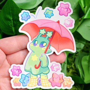 May include: A green creature with a yellow star pattern holds a pink umbrella with a white handle. The creature is wearing yellow and white checkered boots. The background is white with pink, blue, and yellow star-shaped designs.