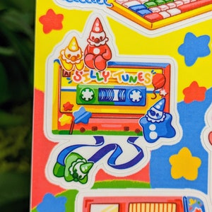 May include: A colorful sticker featuring a cassette tape with the text "Silly Tunes" and cartoon clowns. The sticker is on a yellow background with stars and other colorful designs.