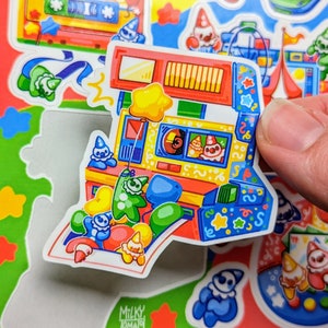 May include: A colorful sticker of a carnival game with a red, yellow, and blue design. The game has a window with a clown inside and a star-shaped balloon floating above. The sticker is surrounded by other stickers of clowns and carnival games.