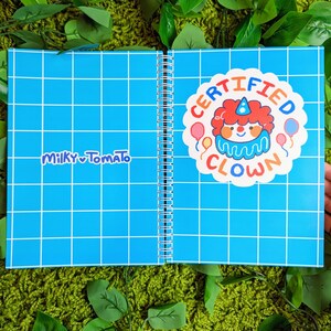 May include: A blue spiral bound notebook with a white grid pattern. The front cover has a sticker that says "CERTIFIED CLOWN" with a cartoon clown illustration. The back cover has the text "MILKY TOMATO" in white.