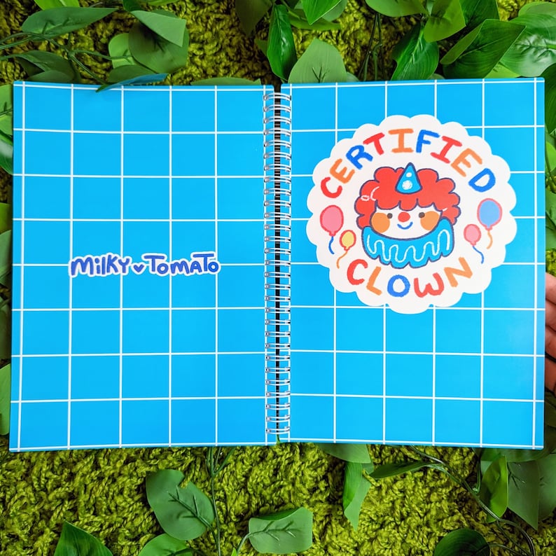 May include: A blue spiral bound notebook with a white grid pattern. The front cover has a sticker that says "CERTIFIED CLOWN" with a cartoon clown illustration. The back cover has the text "MILKY TOMATO" in white.