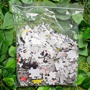 May include: A plastic bag filled with a jumbled pile of colourful jigsaw puzzle pieces. The pieces are mostly white with black, red, yellow, and blue accents.