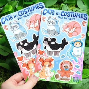 May include: Two sheets of stickers featuring cats dressed in various sea creature costumes. The stickers are on a light blue background with white outlines. The stickers include a cat dressed as a shark, a cat dressed as an octopus, a cat dressed as a crab, and a cat dressed as an orca. The text on the stickers reads "CATS in COSTUMES SEA VERS!!".