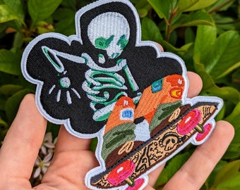 May include: Green skeleton embroidered patch with a black background, riding a skateboard with pink wheels and a brown deck. The skateboard has a detailed design with white, black, and brown lines.