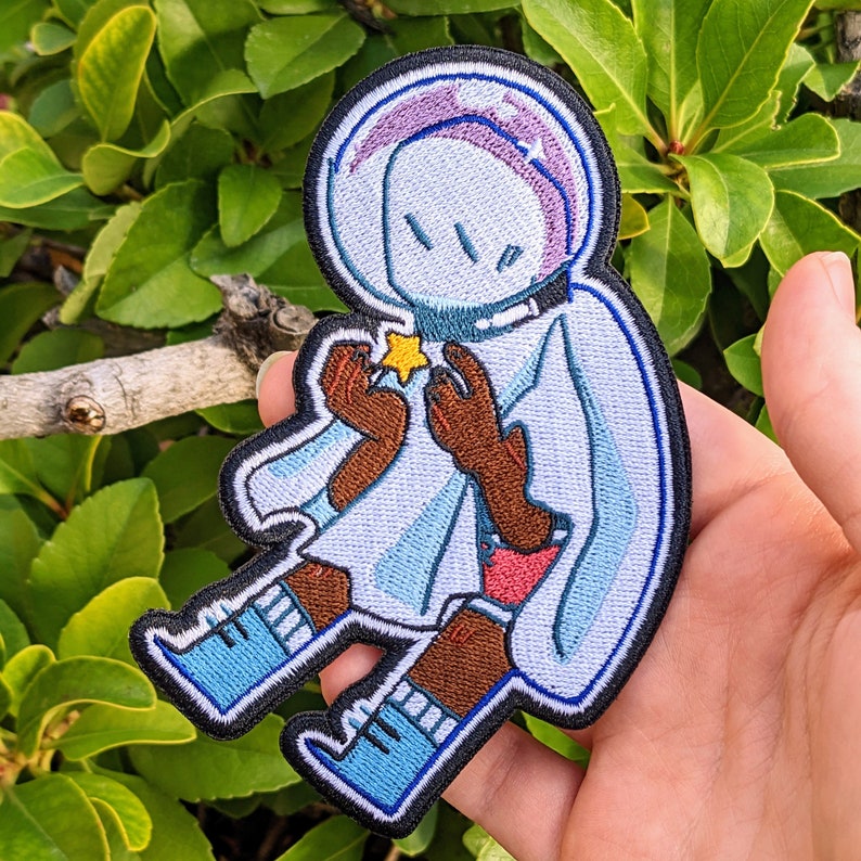 May include: A blue and white embroidered patch of a cartoon character wearing a space helmet and holding a yellow star. The character has brown arms and legs and is wearing blue boots.