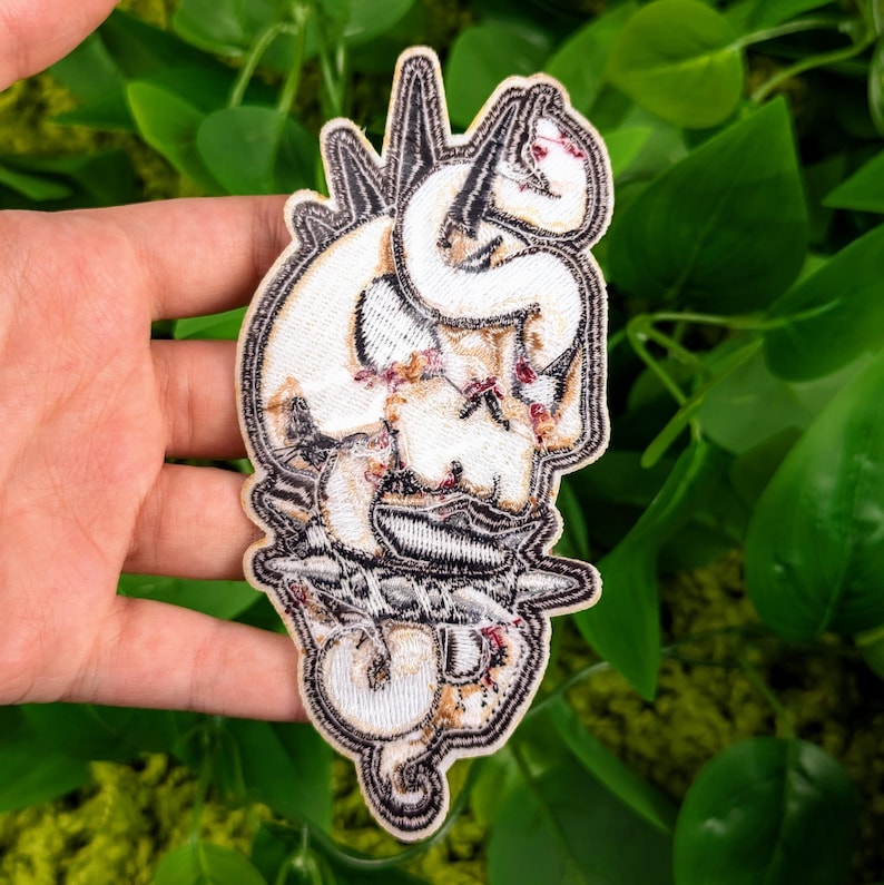 May include: White embroidered patch with a skull and snake design. The skull is surrounded by thorns and the snake is wrapped around the skull's head. The patch has a black outline and is on a white background.