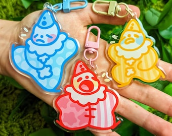 May include: Three colorful keychains featuring cartoon clowns with different expressions. The keychains are made of clear acrylic with a white background and colorful details. The clowns are wearing different colored outfits and have different facial expressions.