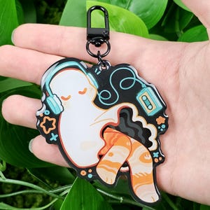 May include: A whimsical keychain featuring a white ghost wearing blue headphones, listening to music from a cassette tape. The ghost has orange and white striped legs. The keychain has a black clasp.