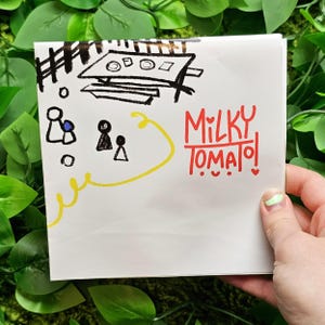 May include: A white card with black and yellow drawings of people and a table. The text "MILKY TOMATO!" is written in red.