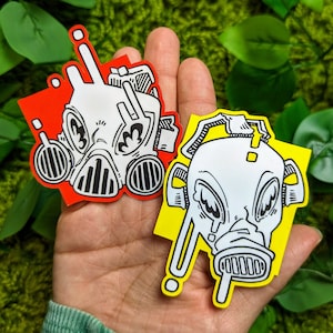 Könnte beinhalten: Zwei schwarz-weiße Gasmasken-Aufkleber mit rotem und gelbem Hintergrund. Die Gasmasken haben einen Comic-Stil und beide tragen einen finsteren Gesichtsausdruck.