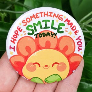 May include: A round button with a smiling cartoon sun design. The sun has a yellow face, orange petals, and green leaves. The button has the text "I HOPE SOMETHING MADE YOU SMILE TODAY!" in green, orange, and red.