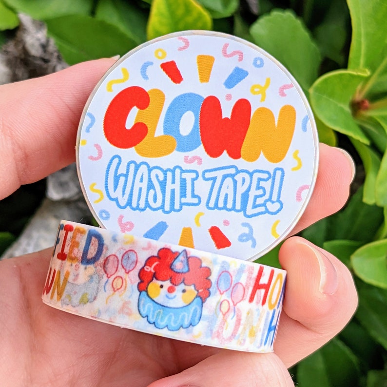 May include: A roll of colourful washi tape with a clown design. The tape features a white background with a clown face, balloons, and the words "CLOWN" and "WASHI TAPE!" The tape is wrapped around a clear plastic spool.