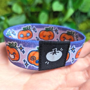 May include: Purple and blue stretchy bracelet with a repeating pattern of orange pumpkins with faces. The bracelet has a black buckle with a white pumpkin on it.