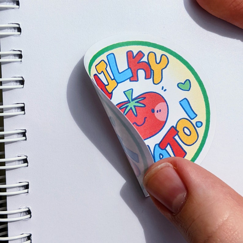 May include: A round sticker with a cartoon tomato design. The sticker has the words "Silky Tomato!" in colorful bubble letters. The tomato has a smiling face and a green stem. The sticker is being peeled back.