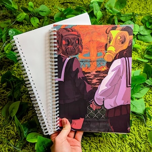 May include: Spiral-bound notebook with a cover featuring an anime-style illustration of two figures wearing gas masks, set against a backdrop of a nuclear explosion. A second, blank notebook is also visible.