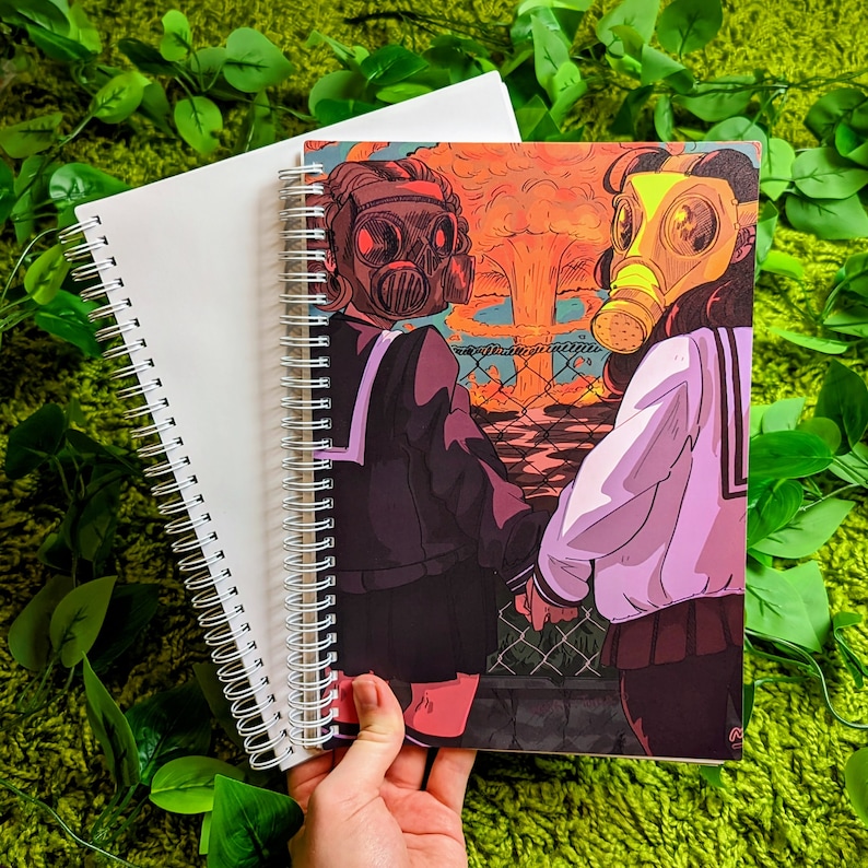 May include: Spiral-bound notebook with a cover featuring an anime-style illustration of two figures wearing gas masks, set against a backdrop of a nuclear explosion. A second, blank notebook is also visible.
