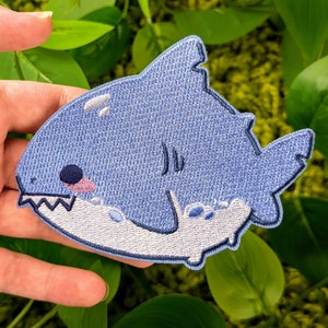 May include: A blue embroidered patch of a cartoon shark with a white mouth and a light blue fin. The shark is smiling and has a small pink tongue.
