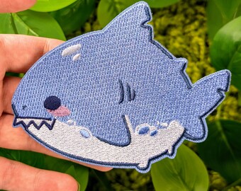 May include: A blue embroidered patch of a cartoon shark with a white mouth and a light blue fin. The shark is smiling and has a small pink tongue.