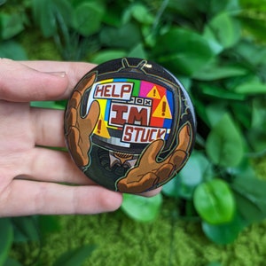 May include: A round button with a cartoon image of a television set with a person's hands sticking out of the screen. The television screen is displaying the words "HELP I'M STUCK" in a colorful, pixelated style.